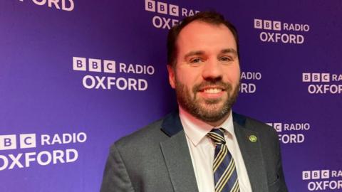 A man in a suit and tie is smiling in front of a BBC Radio Oxford screen.