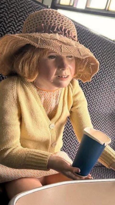 Child mannequin in a yellow cardigan and straw hat sitting on a train seat holding a blue paper cup