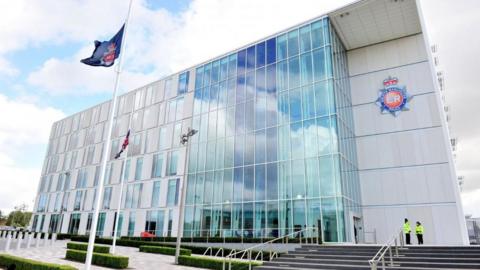 A blue flag with the Greater Manchester police badge is flying at half mast on a flag pole directly in front of the police headquarters. Two police officers are standing to the right-hand side of the building. 