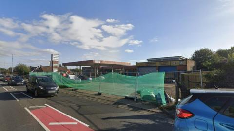 General view of a road with five cars driving towards to right of the frame. In the background there is a see-through green sheet supported by poles and behind it is a closed petrol station