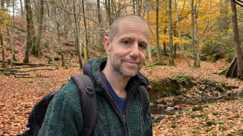 Forager Fergus Drennan stands in a woodland wearing a jumper and small rucksack. He has a shaved head and a stubble beard.