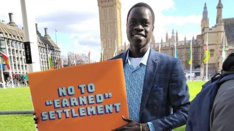 Omot Otor. He is standing outside the Houses of Parliament and is wearing a blue blazer and shirt. He is wearing an orange sign which reads, No to Earned Settlement.