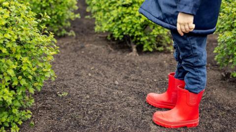 a child in red wellington boots standing on soil, surrounding by plants. Only the legs of the child are visible, in a pair of jeans
