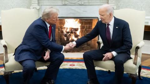 Donald Trump and Joe Biden shake hands in the White House's Oval office. The two are wearing dark suits, white shirts and ties. A fire is burning in a fire place behind them.