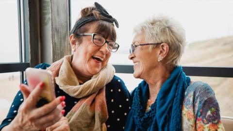 Two female friends sit laughing alongside each other. One is holding her mobile phone in front of them.