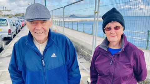 A man wearing a blue-zipped tracksuit top and cap is standing next to his wife, who is wearing a purple coat and dark blue hat. They are on a sea front and looking into the camera.