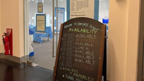 A blackboard leaned up against a glass office door displays the opening times of the Barclays Local branch at Didcot Library. The writing is in different colours and reads: "Welcome to Barclays Didcot".