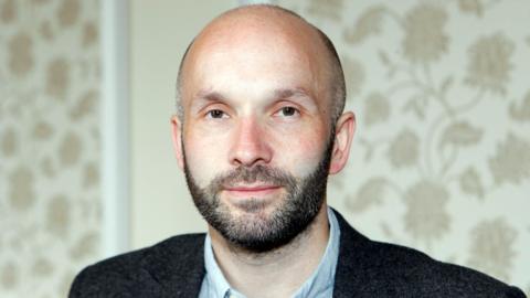 A man in his forties with a shaved head and a short beard. Flowery wallpaper is behind him and he is wearing a suit.