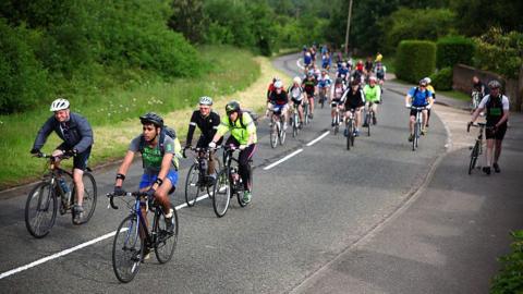 Dozens of cyclists pedal up a hilly road. A couple have dismounted and are walking on the pavement. The road is surrounded by greenery.