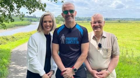 Michael Lilley is standing next to his father John and BBC North West Tonight's weather presenter Kay Crewdson on a path running through a field. He is wearing a T-shirt which says "Prostate Cancer UK".