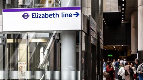 A sign for the Elizabeth line, with purple lettering and branding on a white background, in a busy station. There are travellers and lifts in the background.