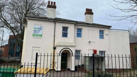 A light-coloured two-storey building with two chimneys. It is behind railings on the photo and a tree is behind the building on the left.