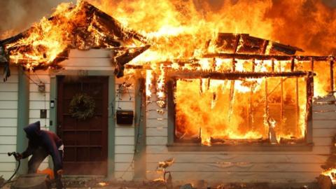 A person with garden hose in front of house that is ablaze