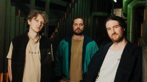 The three members of Bristol band Waldo's Gift face the camera. They are all men, and two of them have beards. The photograph is taken inside a dark room