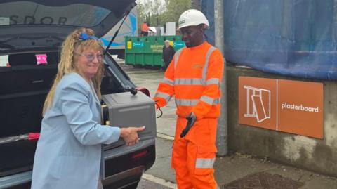 Woman with long light brown curly hair wearing a light blue coat and carrying a grey object which she is handing to a recycling operative in an orange hi-vis and white hard hat. There is a grey car with its boot open behind the woman and a skip covered in a blue tarpaulin behind the recycling operative. It has a red sign which says "plasterboard" in white lettering.