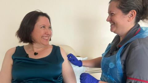 Clare wears a blue, sleeveless top and sits next to a nurse who is rubbing Clare's left arm and is wearing purple medical gloves, ready to vaccinate her