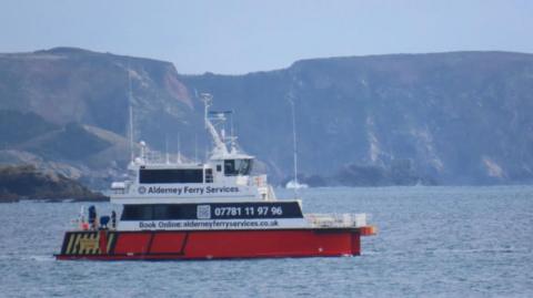 Alderney Ferry Services pictured at sea. The boat is white and red. In the background is an island. Its a hazy day and the sea is relatively calm. 