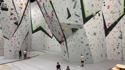An angular climbing walk covering the walls and ceilings of a large building. They are covered in Brightly coloured hand holds and ropes. People stand at the bottom looking at the walls. Crash mats cover the floor.