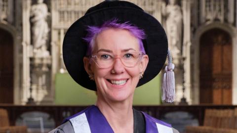 Rebecca Platt is smiling at St Albans Abbey. She has purple hair and is dressed in purple robes which sit n top of her outfit.