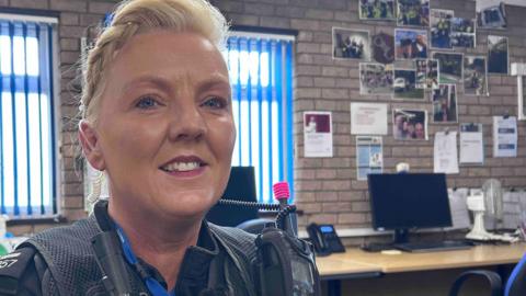 Lorna Montecalvo is sitting inside an office wearing her police uniform. She is looking directly at the camera and smiling and has blonde hair which is braided back. 