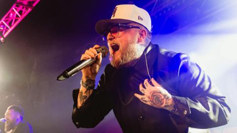 Thom Rylance is wearing a white cap, orange tinged glasses and black jacket. He singing into a microphone on stage.
