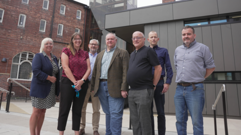 Simkins is pictured with six other staff from the adults education facility and library. They are stood outside outside the new library section and adult education building. There are two women and six men.