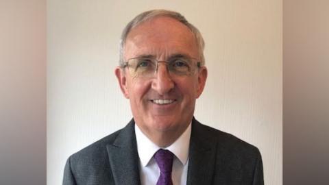 A man with short grey hair and glasses smiles at the camera. He is wearing a dark grey jacket, white shirt and purple tie. 