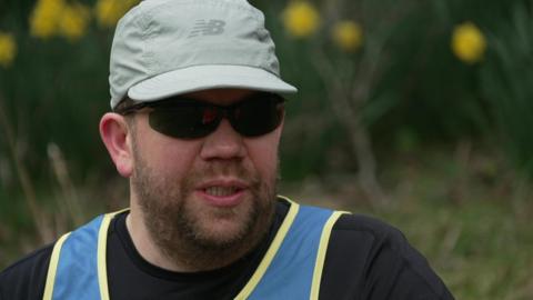 A man in a cap and sunglasses, wearing a black t-shirt and blue and yellow vest. 