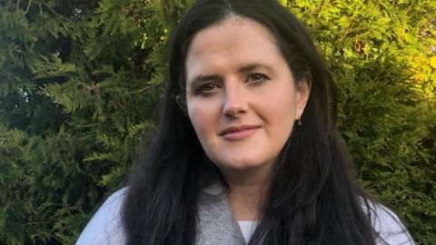 Claire Sugden, who has black hair and brown eyes, wearing a grey scarf and white top. She looks into the camera and is standing in front of a green hedge.