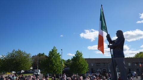 A crowd gathered around a statue of Bobby Sands, with an Irish flag hanging on it. The sky is clear and blue above. There are some trees and a row of houses in the background. 