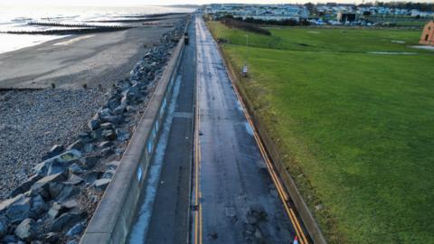 A bird's eye view of a road and a path. The seafront is on the left and a grass field is on the right.
