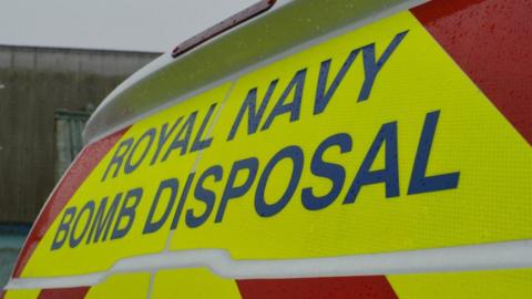 The back of a white van which has yellow and red chevron and a sign saying "Royal Navy Bomb Disposal"