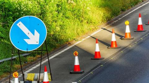 Roadworks block one lane of a carriageway. A circular blue sign with a white arrow directs cars around a line of cones.