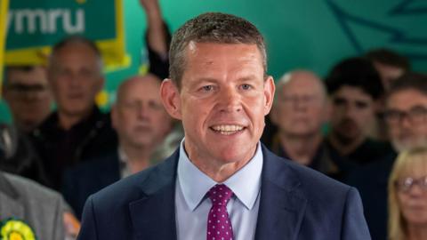 Leader of Plaid Cymru Rhun ap Iorwerth speaking after Plaid Cymru's Senned election victory. He is wearing a navy-blue blazer, light blue shirt, and a purple polkadot tie. He is smiling and looking at the camera. People can be seen stood behind him.