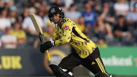 Kamran Dhariwal holds a bat in two bands to his right as he runs on the pitch during a T20 game for Gloucestershire
