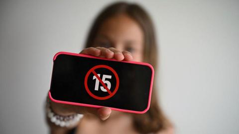 A teenager holds her smartphone displaying a logo "ban under 15". The phone screen is black with a crossed out 15 in the middle of it. The person is blurred in the background and the phone is held in front of them facing the camera.