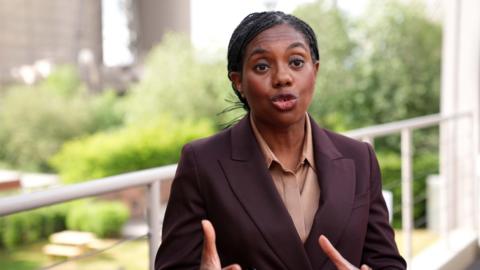 A woman wearing a claret coloured suit jacket and orange shirt is standing on a balcony.