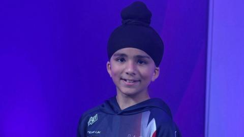 A young boy smiling at the camera. he is wearing a turban and a sports hooded top and in a room with purple walls.
