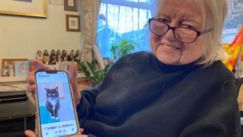 A woman with mid-length white hair and glasses, wearing a black jumper, shows a photograph on her phone of a black cat with white paws and a white chest.