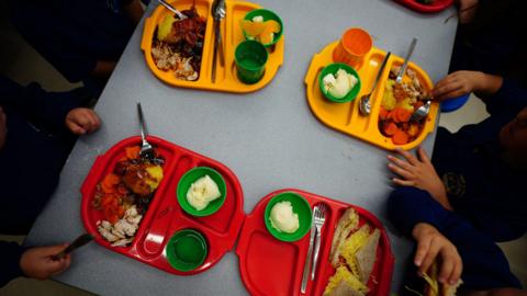 Children sit at a table and eat from four lunch trays which are filled with food. Two of the trays are red and two of them are yellow.