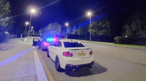 The seized white BMW M2 on the street and a police car in front of it. It's night time.