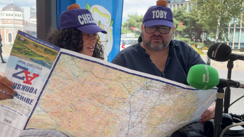 A man and a woman stare at a large map of Yorkshire. They are wearing hats with their names on: Chelsey and Toby.