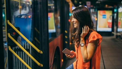 A young woman is about to step onto a bus. It is night time in a city. The bus door is open. She is has shoulder-length dark hair and is wearing a short-sleeved orange top and jeans. She has her mobile phone in her right hand and is smiling. An illuminated advertisement posted attached to a bus stop can be seen in the background as well as other city lights.