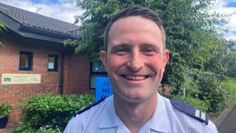 Peter Bradley in his NIFRS uniform, a white shirt with black shoulder pins, smiling
Pictured in front of a red brick building with a large green bush beside it