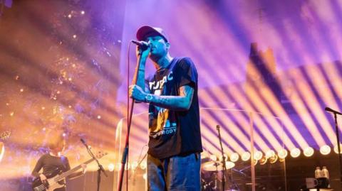 A singer on stage, with a bassist to his right. He is wearing jeans, a black printed t-shirt and cap. Tattoos can be seen on his bare arms and the stage is lit with purple and yellow lights.