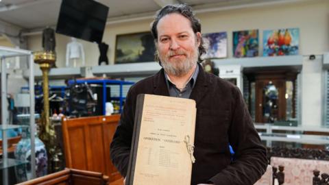 Matt Crowson holds a rare top-secret blueprint of the D-Day landings. He is wearing a brown jacket and has a grey beard. He is holding a dated looking bundle of papers with "Operation Overlord" visibile on the front.