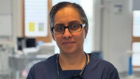 Dr Kainth is looking at the camera ans smiling. She is wearing scrubs and a stethoscope around her neck. She is wearing glasses and had dark hair tied back.