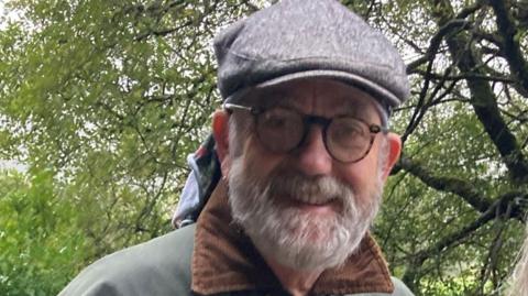 A man with a beard and glasses, wearing a grey flatcap