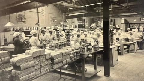 An old photo, in black and white. It shows a group of about 30 factory workers all wearing white bonnets and white aprons. They are all gathered around different tables which have clear glass jars of sweets on them. There are also lots of parcels at the forefront of the photo.