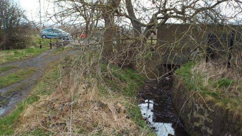 The tiny stream at Cut Throat Dene in Sunderland. The water is murky with leaves floating in it. A tree is above the stream, which is next to a path. A blue car is parked at the top of the stream.

